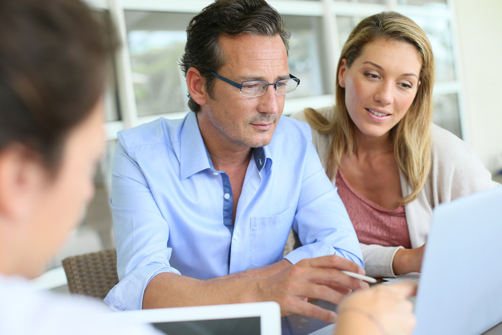 A group of diverse sales professionals, men and women of various ethnicities, are gathered around a laptop in a meeting room. They are all focused on the laptop screen, with some leaning in closer and others nodding in agreement. The atmosphere seems engaged and collaborative.